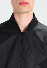 Svart bomberjacka med en slät, glänsande yta. Har en ribbad krage och en central dragkedja, vilket ger en figursydd look.