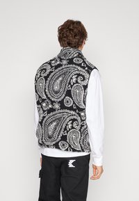 Gilet senza maniche con base nera e motivo paisley bianco. Realizzato in tessuto testurizzato, con colletto alto e orlo leggermente curvato.