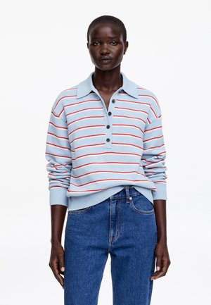 Femme portant un polo à manches longues bleu clair avec des rayures horizontales rouges et blanches, et un jean bleu taille haute, debout face à l'avant.