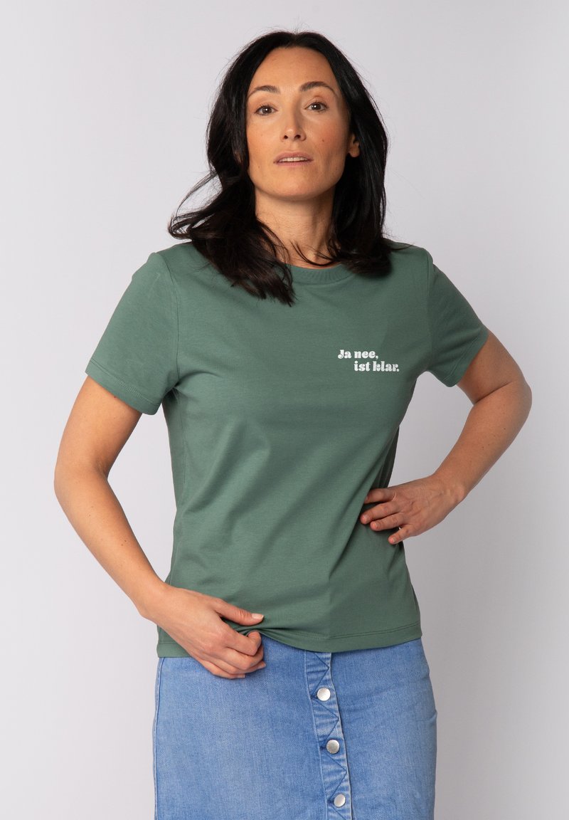 Grünes Baumwoll-T-Shirt mit kurzen Ärmeln, bedruckt mit dem weißen Text "Ja nee, ist klar." Modell kombiniert mit einem Jeansrock, die Hände in die Hüften gestützt.