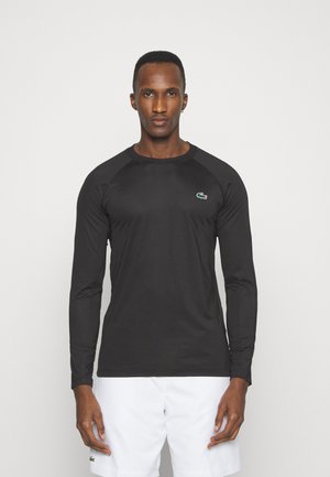 Chemise noire à manches longues en tissu lisse, avec des manches raglan et un petit logo vert sur la poitrine.
