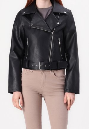 Frau trägt schwarze Leder-Bikerjacke mit Reißverschlusstaschen und Gürtel, kombiniert mit beigen Hosen, vor einfarbigem Hintergrund stehend.