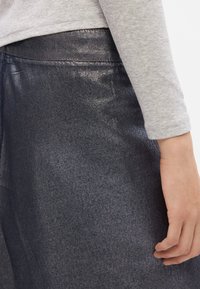Denim sijonas su blizgiu, tamsiai mėlynu paviršiumi. Turi lygų tekstūrą ir paprastą juosmenį. Rankos padėta ant sijono.