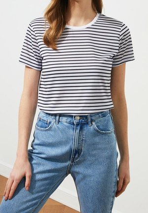 Frau trägt ein weißes Kurzarm-T-Shirt mit schwarzen horizontalen Streifen, das in hoch taillierte blaue Jeans gesteckt ist, und steht drinnen.