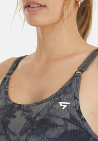 Grijze tie-dye sporttop met ribbeltextuur, verstelbare bandjes en een wit logo aan de linkerkant. Close-up van de halslijn en schoudergebied.
