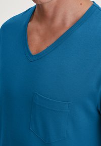 T-shirt en coton bleu pour homme avec un design à col en V, comportant une petite poche sur la poitrine gauche. Texture lisse, d'épaisseur moyenne.