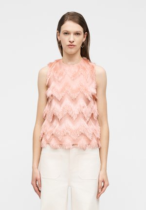 Vrouw draagt een mouwloze roze top met een getextureerd zigzagfranje en witte broek met hoge taille, staand tegen een effen achtergrond.