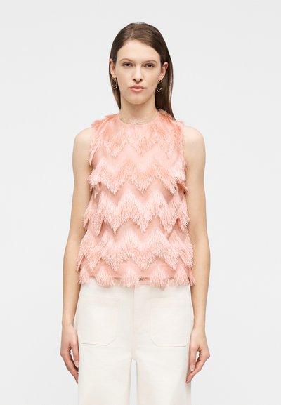 Femme portant un haut rose texturé sans manches avec des franges en zigzag et un pantalon blanc taille haute, debout devant un fond uni.