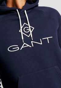 Felpa con cappuccio blu navy con cordini bianchi. Presenta un logo a diamante con la lettera "G" e il testo "GANT" ricamati in lettere bianche.
