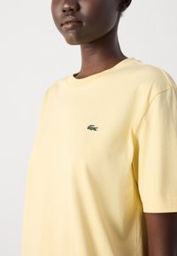 Gul bomullströja med rund halsringning, lös passform och en broderad grön krokodil-logotyp på bröstet. Slät yta, korta ärmar.