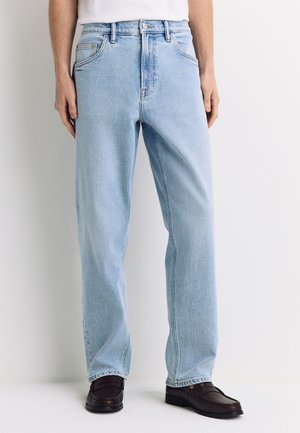 Person iført lyseblå jeans med lige ben og mørkebrune slip-on sko, stående mod en ensfarvet hvid baggrund.