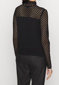 Pull noir avec un design en maille présentant de grandes découpes rondes sur les manches et le dos, un panneau uni sur le bas du corps, et un ourlet côtelé.