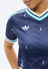 Detail osoby v tmavě modrém sportovním tričku Adidas s světle modrým a bílým lemováním a vzorovanou strukturou, s logem Adidas na hrudi.