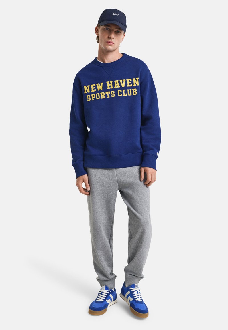 Marineblå sweatshirt med gult "NEW HAVEN SPORTS CLUB" tekst, parret med grå sweatpants og blå sneakers med hvide accenter.
