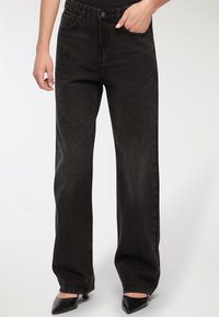 Szerokie czarne dżinsy o gładkiej fakturze, z zapięciem na guzik i przednimi kieszeniami. Noszone z czarnymi szpilkami ze spiczastym noskiem.