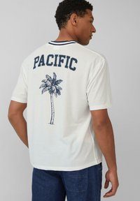 Witte T-shirt met korte mouwen met "PACIFIC" in marineblauw op de achterkant, met een palmboomafbeelding onder de tekst. Ronde hals.