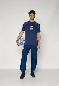 Jersey atlético azul marino con acentos en rojo y blanco, que presenta un logo de Adidas y un distintivo del equipo, combinada con pantalones de chándal a juego en azul marino y zapatillas negras.