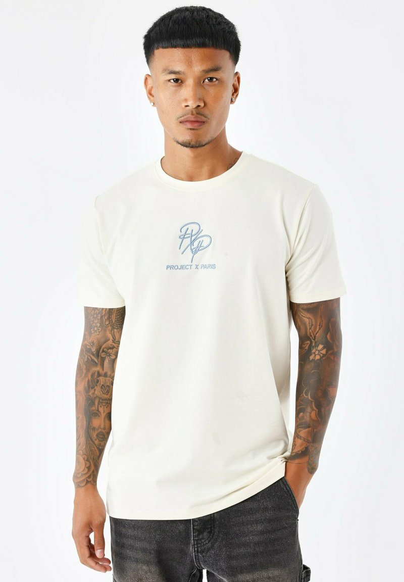 T-shirt en coton crème à manches courtes avec un logo bleu "PROJECT X PARIS" sur la poitrine. Le modèle porte un jean foncé ; des tatouages visibles sur les bras avec des designs intricats.