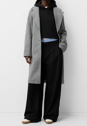 Personne portant un long manteau gris par-dessus un sweat à capuche noir, un pantalon large noir, une chemise rayée bleue qui dépasse, et des chaussures blanches avec des semelles brunes.