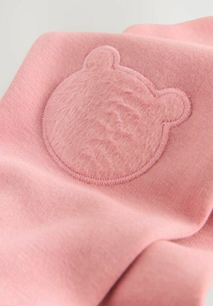 Tissu rose avec une applique ronde en forme de tête d'ours texturée. L'applique a une surface douce et duveteuse, avec des coutures visibles autour des bords.