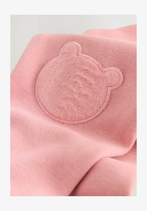 Ružičasta tkanina s okruglim, teksturiranim zakrpom u obliku medvjedove glave. Zakrpa ima mekanu, pahuljastu površinu s vidljivim šavovima oko rubova.