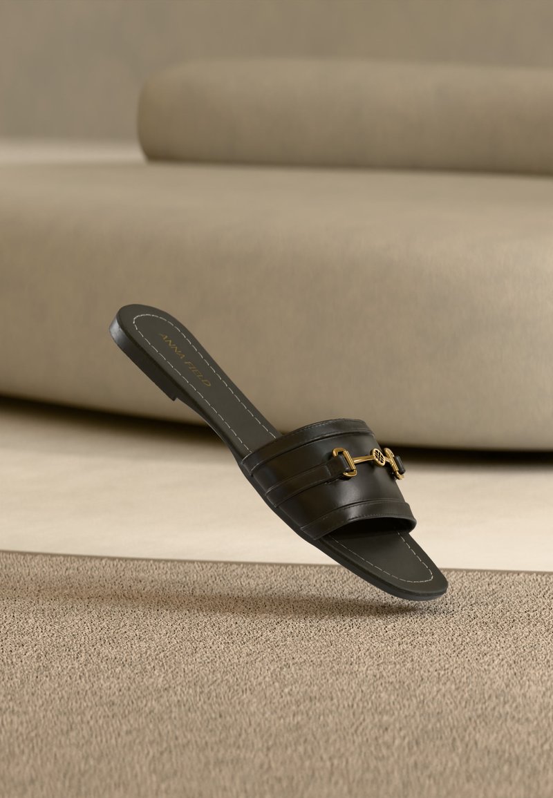 Sandale noire en cuir à enfiler avec détail de boucle dorée posée sur un tapis beige devant un canapé rembourré couleur tabac.