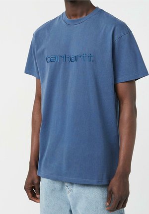 Man wearing a faded blue Carhartt t-shirt with short sleeves and light blue denim jeans, standing against a plain background.