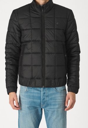 Homme portant une veste matelassée noire à fermeture éclair avec des poches latérales et un jean bleu clair, debout devant un fond uni.