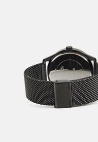 Black mesh strap watch with a round case, shiny black dial, and clasp featuring the "Guess" logo engraved on the side.