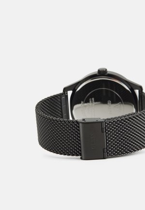 Schwarzes Uhrenarmband aus Mesh mit einem polierten, runden Gehäuse. Die Rückseite zeigt eine reflektierende Oberfläche mit der Gravur "GUESS" in der Nähe der Schließe.