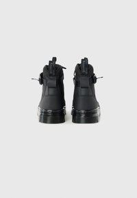 Un par de botas negras hasta el tobillo con suelas resistentes, lengüetas traseras que dicen "CON SUELAS QUE REBOTAN" y correas con hebillas en los lados sobre un fondo blanco.