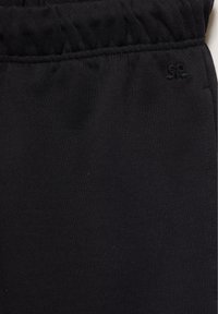 Gros plan sur un tissu noir avec une ceinture élastique froncée et un petit logo brodé sur le côté droit.