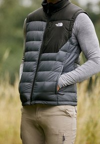 Graue und schwarze Steppweste mit hohem Kragen, Reißverschluss vorne und Seitentaschen; getragen über einem langärmligen grauen Hemd im Freien.