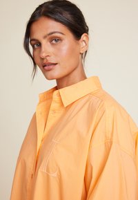 Kortermet skjorte med krage og knapper i klart oransje bomull. Har et brodert bokstav "L" på brystet og en avslappet passform.