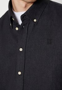 Primo piano di una persona che indossa una camicia a maniche lunghe grigia scura con bottoni sopra una maglietta bianca, con un piccolo logo ricamato a doppia linea sul petto.
