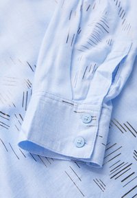 Himmelblaues Hemd aus strukturiertem Stoff, mit einem Muster aus feinen, dunklen Linien. Die Manschette hat zwei himmelblaue Knöpfe und eine saubere Verarbeitung.