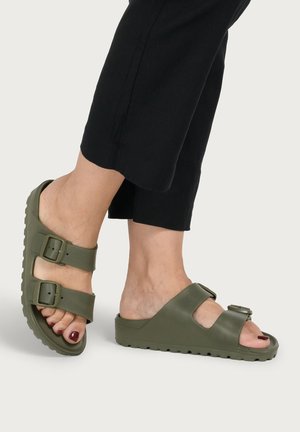 Feet wearing olive green slip-on sandals with two adjustable straps and black cropped pants on a plain background.