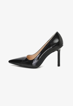 Pamelaige en cuir verni noir à talons hauts avec un bout pointu et une découpe sur le côté, présentée avec un talon fin pour accentuer la hauteur.
