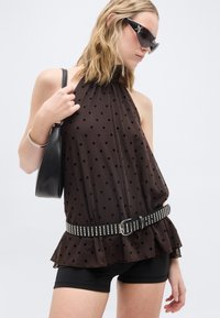 Γυναίκα που φορά διαφανές μαύρο τοπ πουά με λουράκι στο λαιμό και ζώνη με τρουκς, μαύρο σορτς, γυαλιά ηλίου με αστέρια, ασημένιο βραχιόλι και κρατά μαύρη τσάντα.