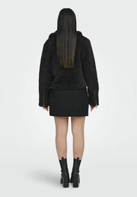Giacca nera e pelosa con colletto alto, maniche con zip, abbinata a una mini gonna nera e stivali con tacco spesso.
