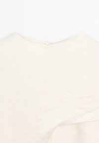 Lichtbeige kledingstuk van een zachte, gestructureerde stof. Heeft een ronde halslijn en een klein knoopdetail op de achterkant.