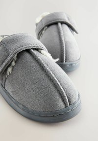 Des chaussons en daim gris avec une douce doublure blanche, dotés d'une sangle à crochet et boucle pour un ajustement sécurisé et des accents cousus le long des coutures.