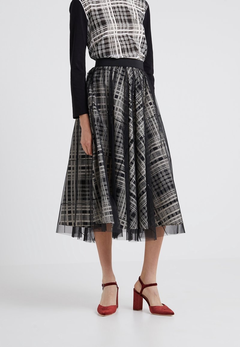 Femme portant une jupe midi noire et blanche à motifs, un haut noir à manches longues et des chaussures rouges à talons hauts, debout sur un fond uni.