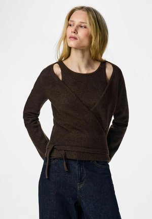 Blondynka w ciemnobrązowym, długim swetrze typu wrap z wycięciami na ramionach i ciemnoniebieskich dżinsach, patrząca w bok.