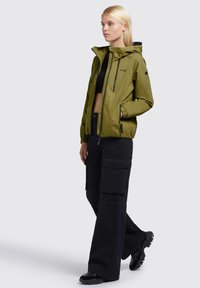 Olive green waterproof jacket with a hood, elastic cuffs, and zipped pockets, paired with loose black cargo pants and black footwear.