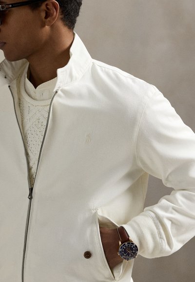 Vyras vilki baltą striukę su užtrauktuku virš tekstūruoto megztinio, rudas laikrodis su odiniu dirželiu, ranka kišenėje, žvelgia į kairę.