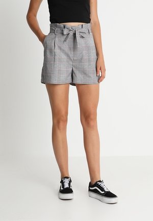 Femme portant un short taille haute à carreaux gris avec une ceinture nouée, un crop top noir et des baskets noires et blanches, debout sur un fond blanc.