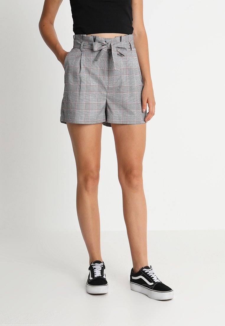 Femme portant un short taille haute à carreaux gris avec une ceinture nouée, un crop top noir et des baskets noires et blanches, debout sur un fond blanc.