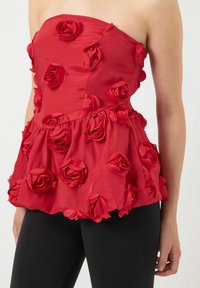 Rød stropløs top lavet af satin, prydet med stofroser, med en peplum design og glat tekstur. Båret sammen med sorte leggings.