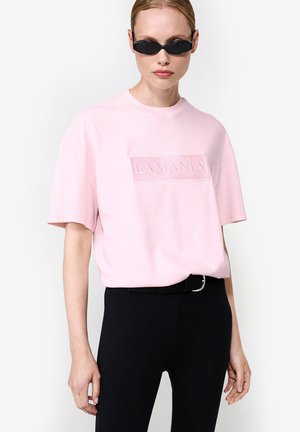 Kobieta ubrana w czarne wąskie spodnie z paskiem, jasnoróżowy oversize T-shirt z napisem "LAMANIA" oraz wąskie czarne okulary przeciwsłoneczne na białym tle.